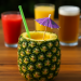 Pineapple drink with purple umbrella, on a table with other drinks.